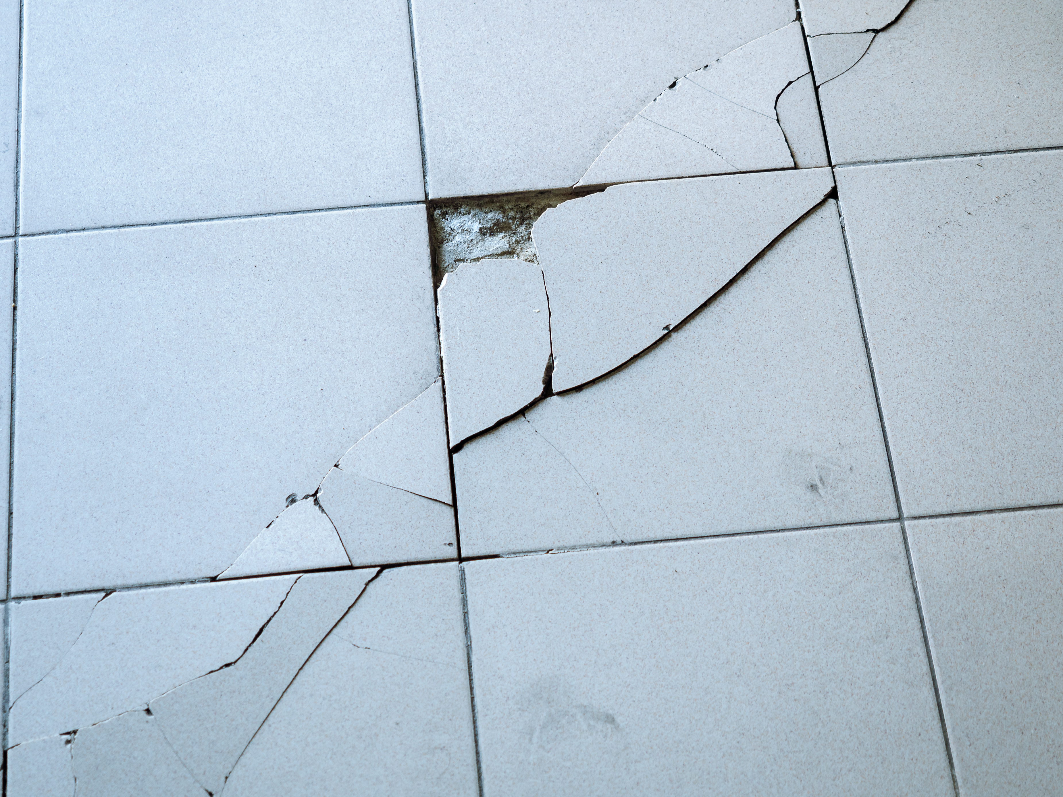 Close-up photo of light gray tile with large diagonal cracks and chips.