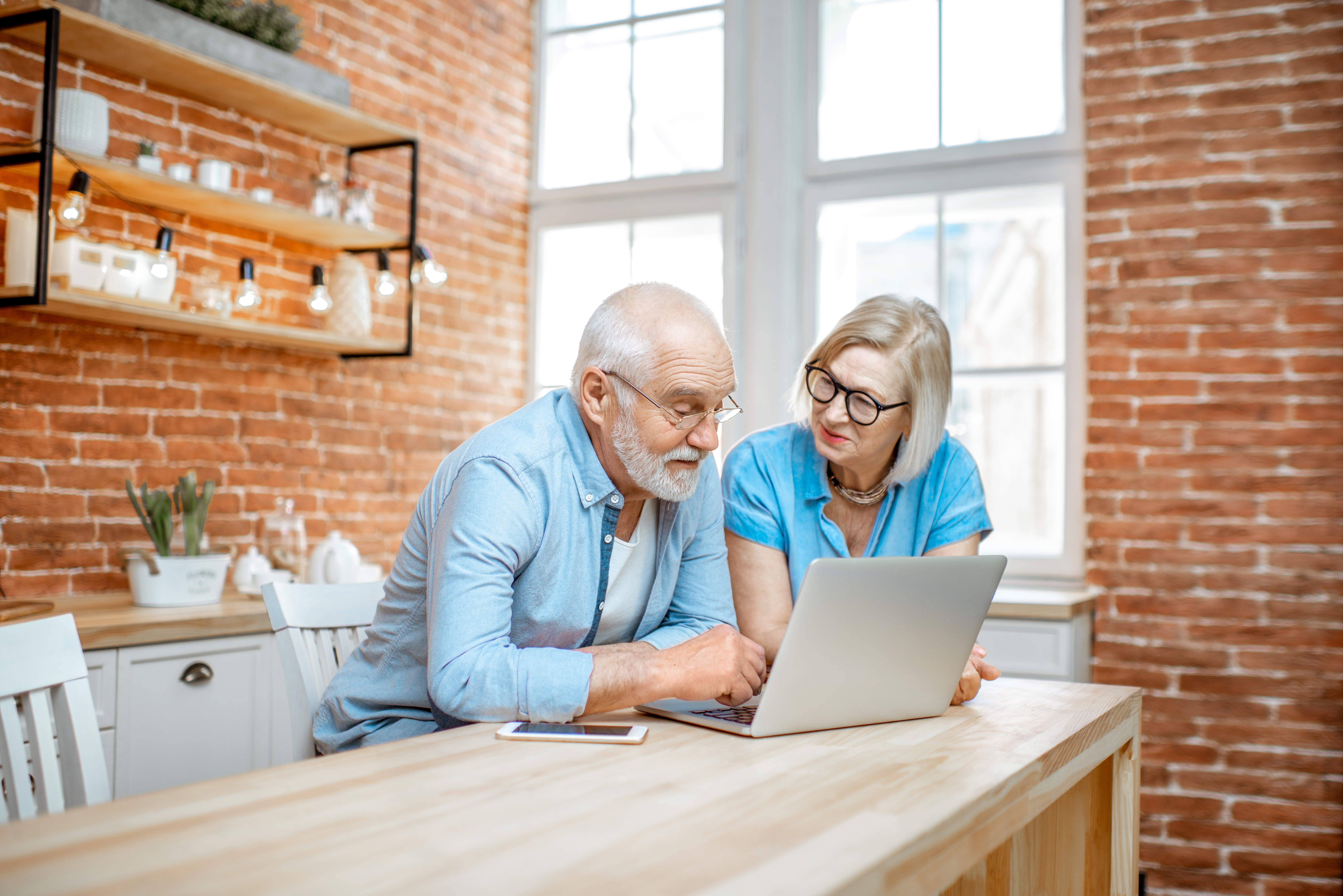 Beautiful senior couple in blue shirts sitting together with laptop on the kitchen at home